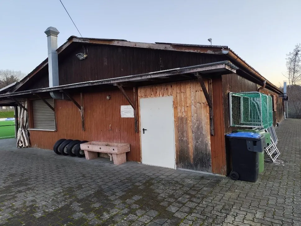 Das heutige Clubhaus von außen: verwitterte Holzfassade, verzogene Dachkante, davor gestapelte Reifen und Mülltonnen